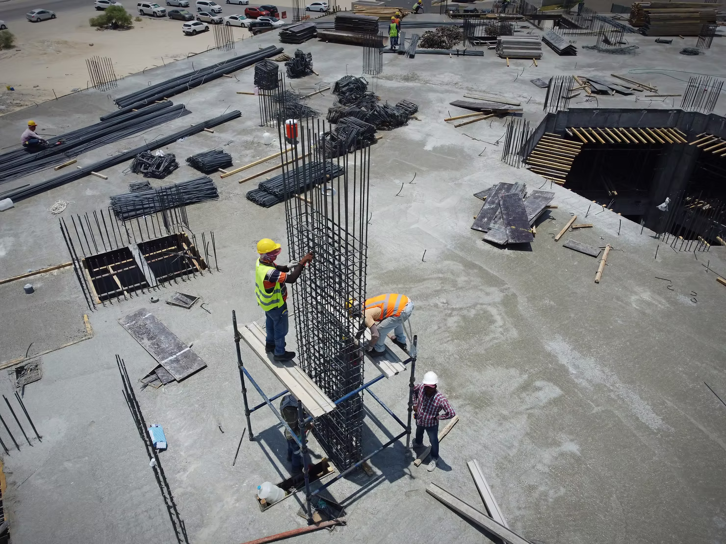 Aerial view of a concrete deck on an active construction site, with workers on scaffolding tying a tall rebar column cage