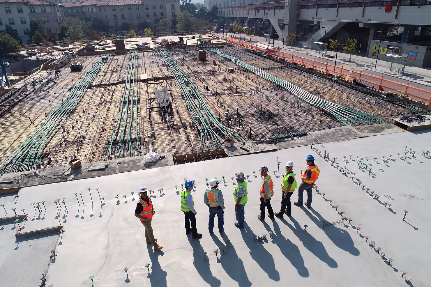 Construction crew in hard hats and safety vests standing on a concrete deck overlooking rebar and post-tension cables on a large building site