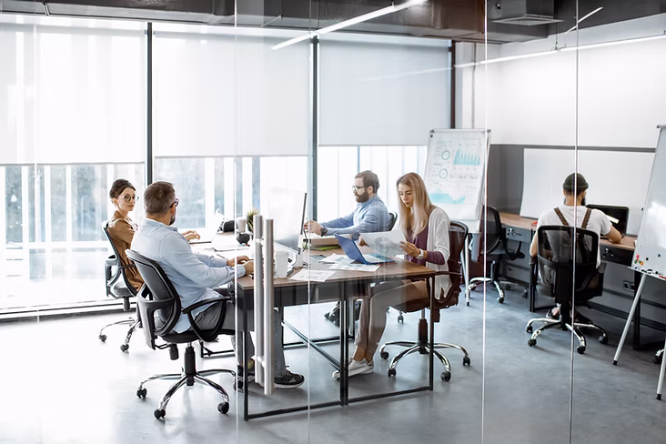 Team members reviewing documents and laptops around a shared table in a glass-walled modern office