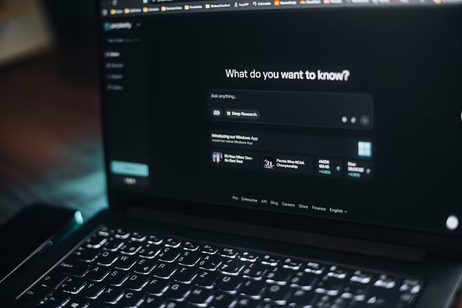Laptop in a dim room showing an AI search assistant asking “What do you want to know?” above a backlit keyboard