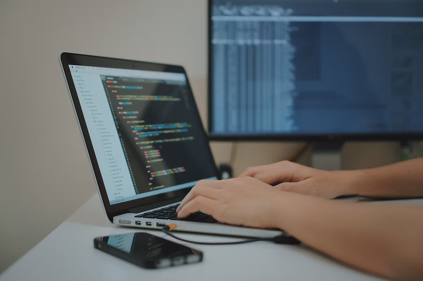 Hands typing on a laptop showing a code editor, with a second monitor of data and a smartphone on a white desk