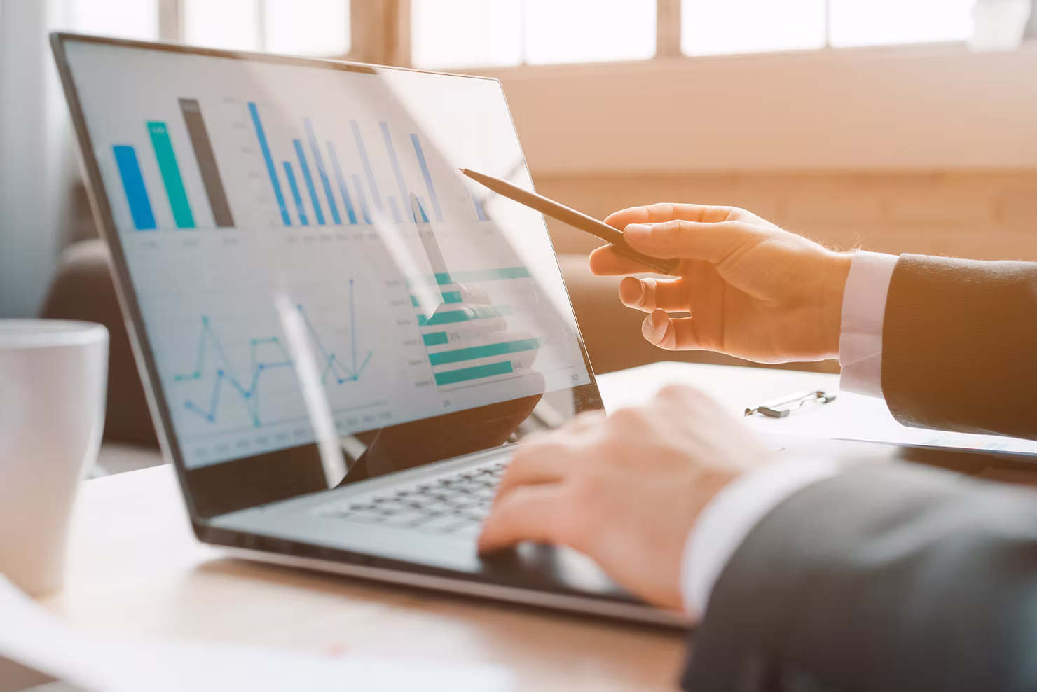 Businessman pointing a pen at bar and line charts on a laptop screen while reviewing project data in a sunlit office