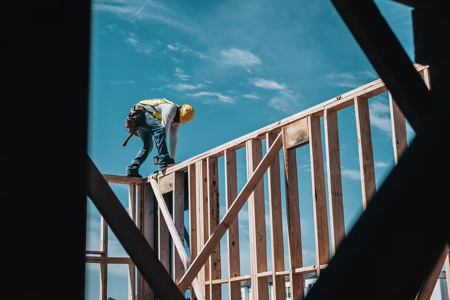 Construction worker in a hard hat and safety vest balancing on top of a wood-framed wall against a blue sky