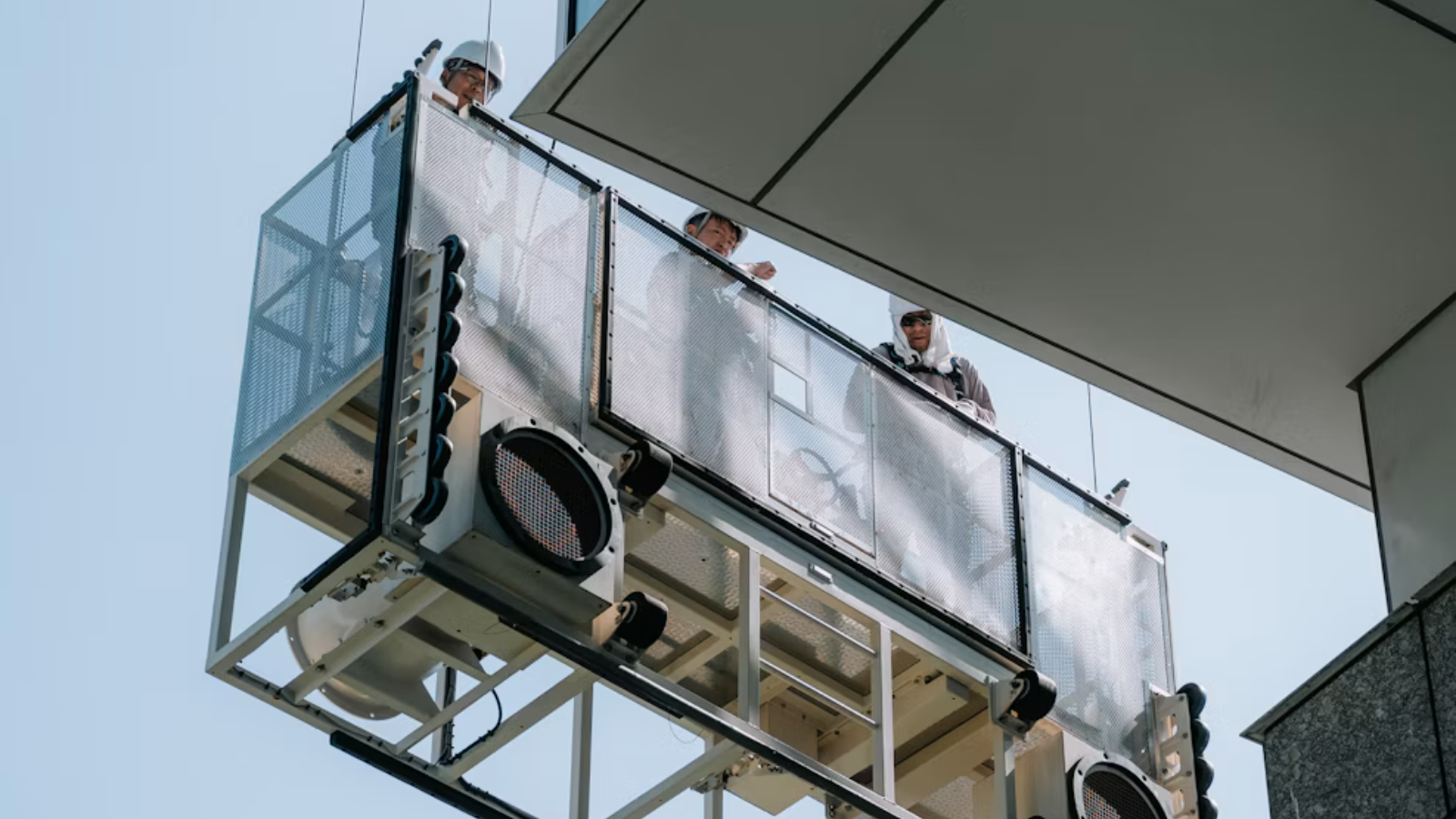 Three workers in hard hats on a suspended maintenance platform hanging beside a glass office building against a clear sky