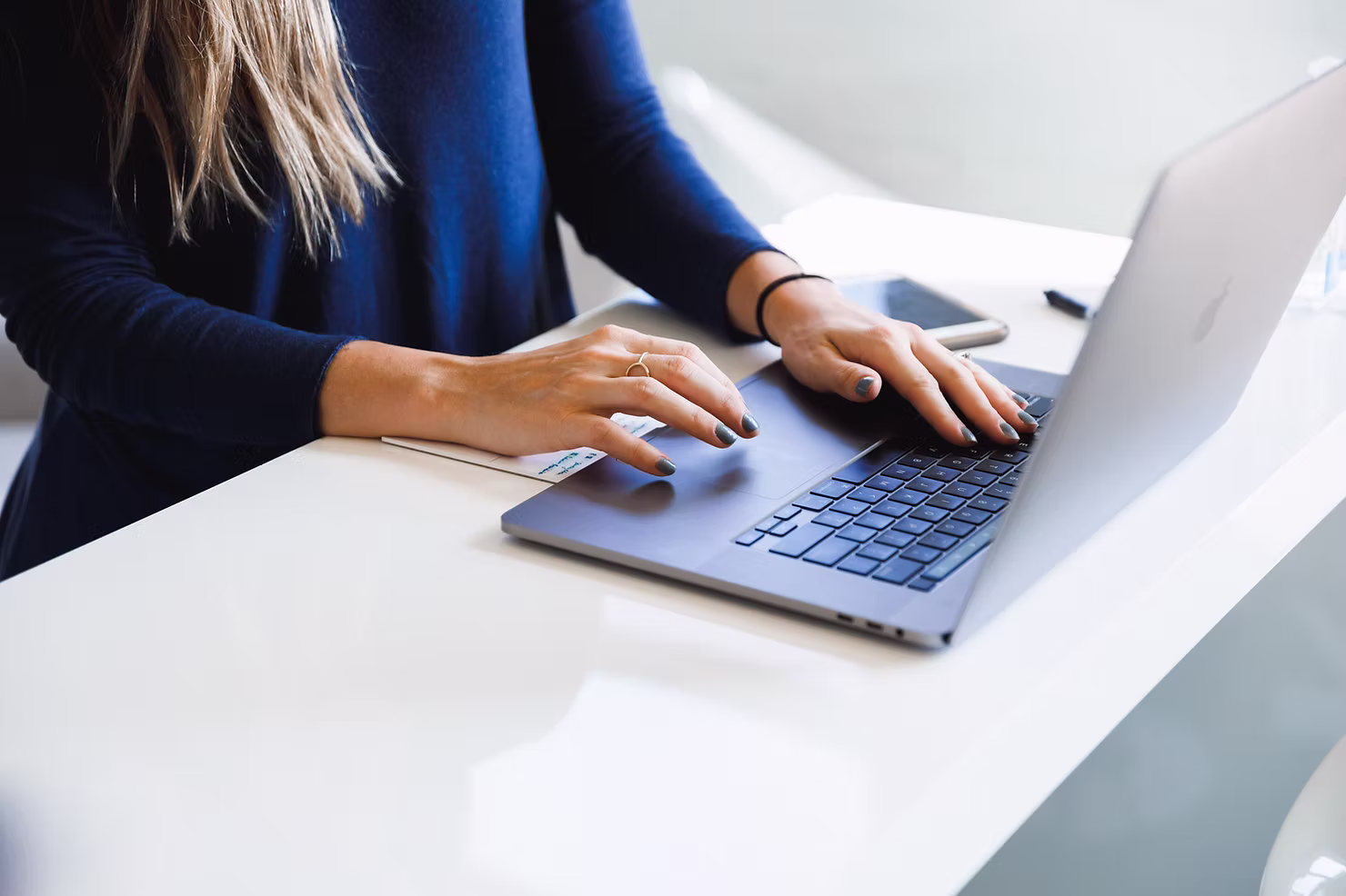 Close-up of a woman in a navy sweater typing on a silver laptop at a bright white desk