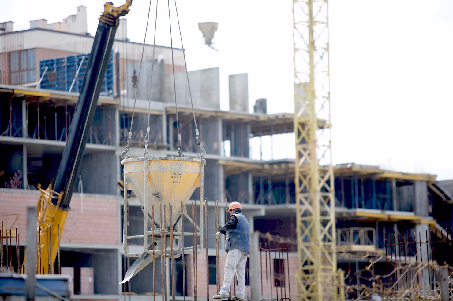 Construction worker in a hard hat guiding a crane-suspended concrete bucket over rebar at a multi-story building site