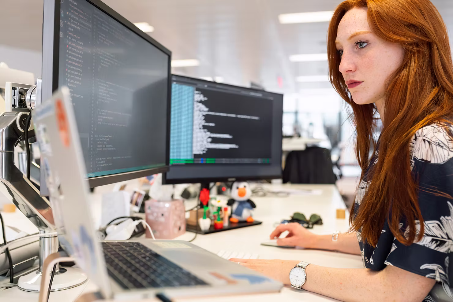 Woman with long red hair focused on code and data on two desktop monitors and a laptop in a bright open-plan office