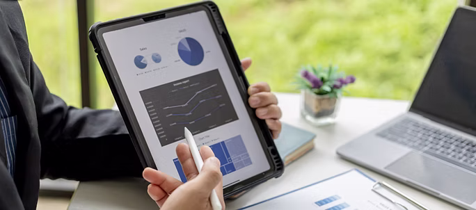 Professional holding a tablet with a stylus, reviewing pie charts and a business report dashboard beside a laptop