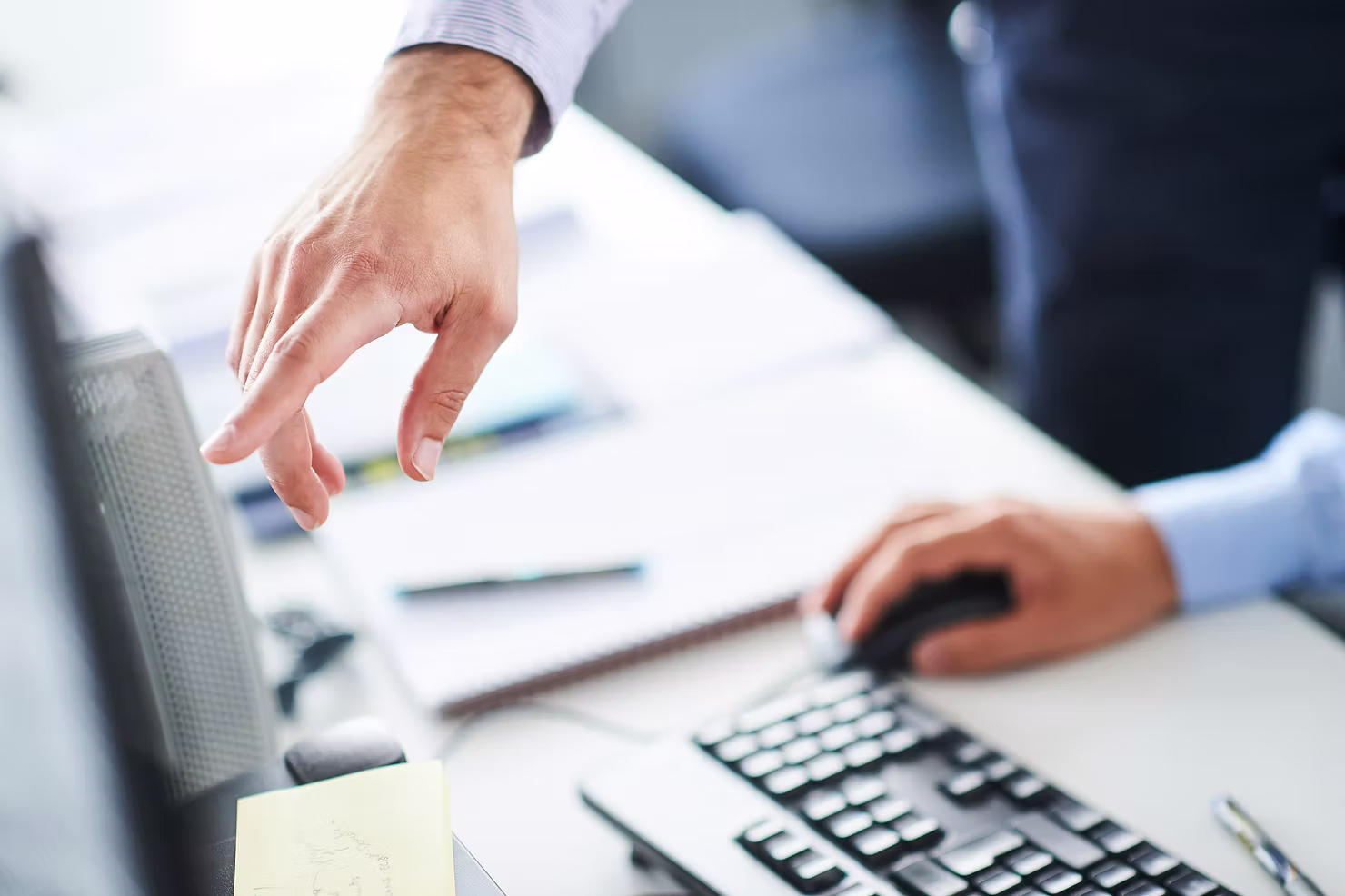 Two colleagues at an office desk, one pointing at a computer monitor while the other uses the mouse and keyboard