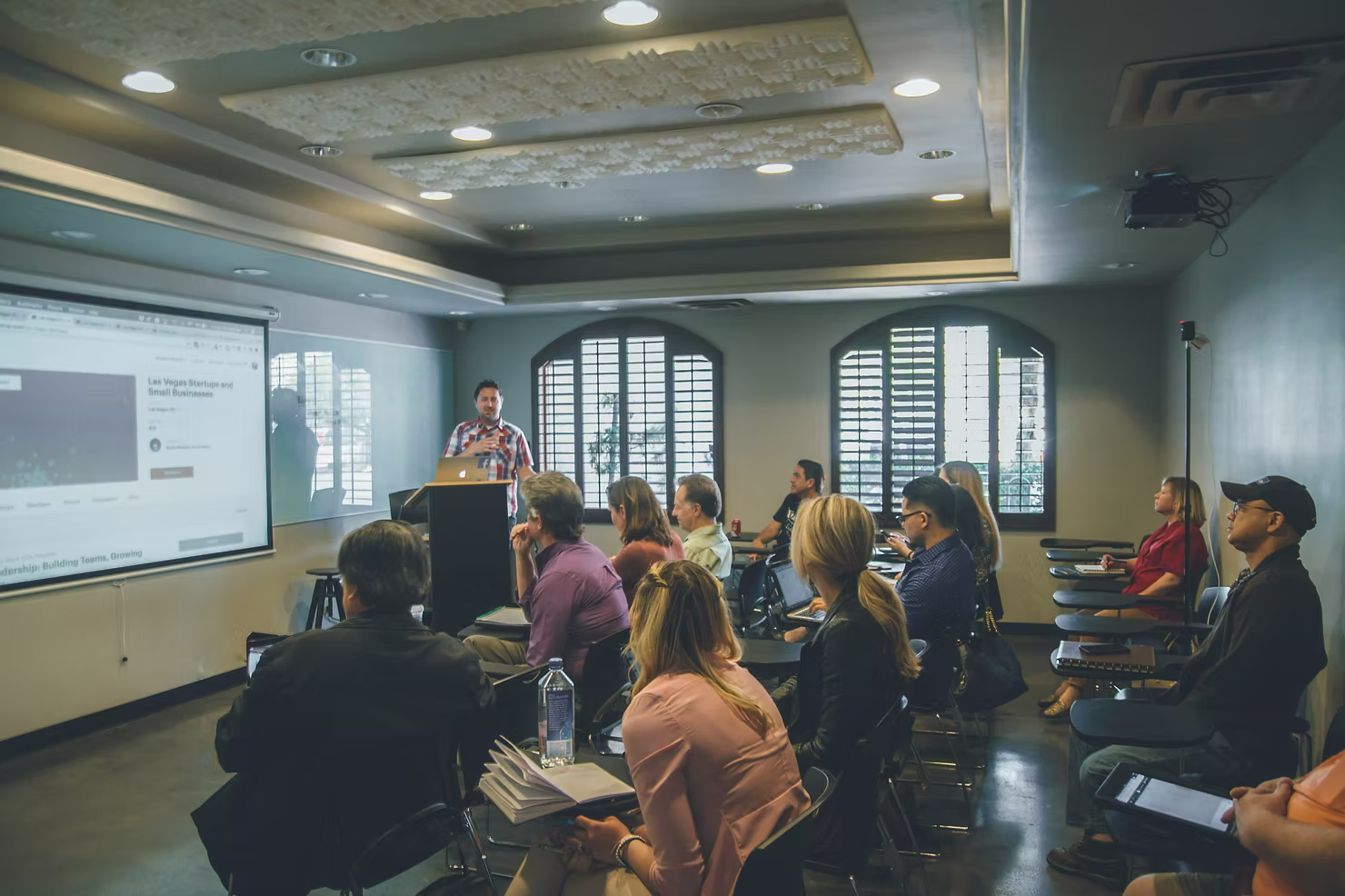 Presenter at a podium leading a workshop as a room of professionals with laptops and notebooks watches a projected screen