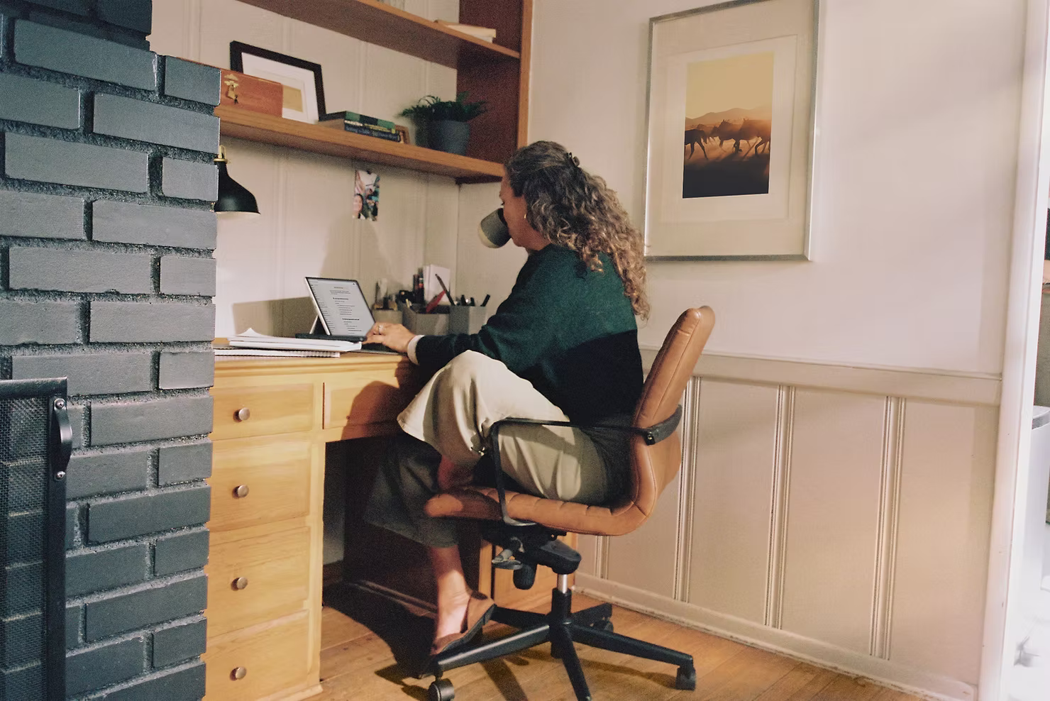 Woman with curly hair sipping coffee while working on a tablet with a keyboard at a wooden desk in a home office
