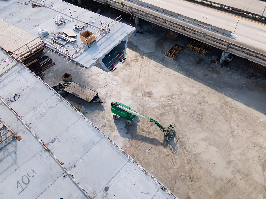 Aerial view of a highway bridge construction site with precast concrete deck segments, stacked formwork, and a green boom lift