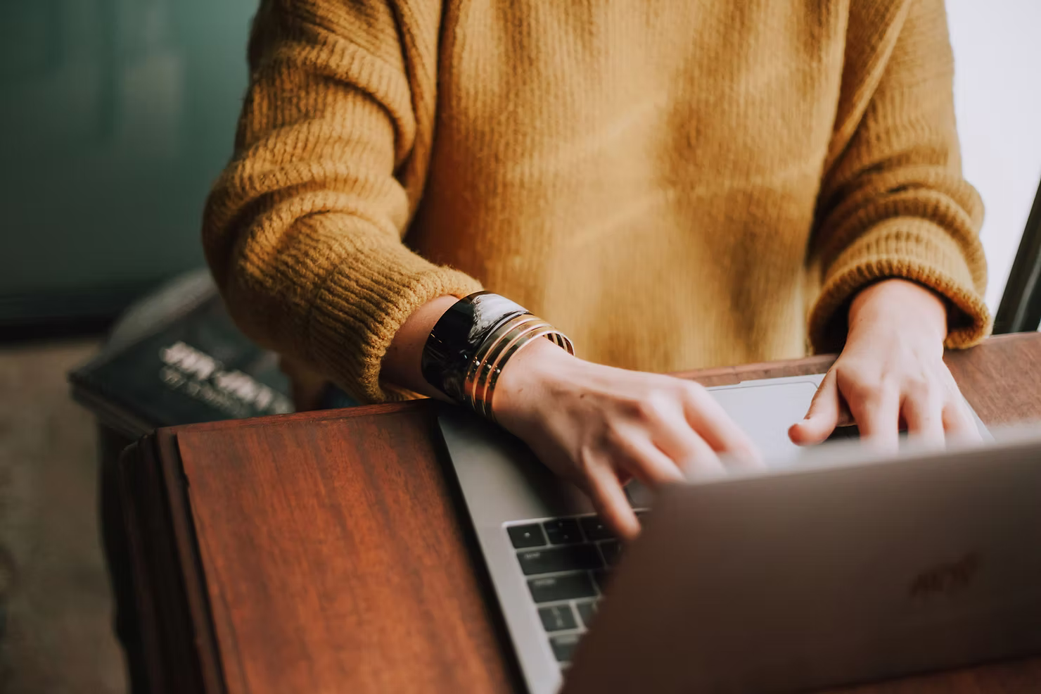 Close-up of hands typing on a laptop at a wooden desk, a person in a mustard knit sweater wearing gold and black bangles